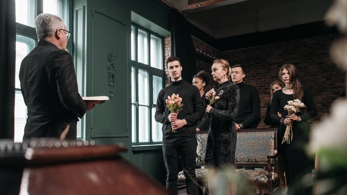 A solemn indoor ceremony with people holding flowers and listening to a speaker.